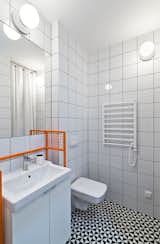 The bathroom features graphic eye-catching Vives floor tiles surrounded by Opoczno wall tiles and clean white plumbing fixtures. The custom-designed orange sink frame adds a whimsical pop of color.