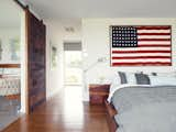 He built the walnut bed and nightstands in the master bedroom with the help of Hannah’s two brothers. One of his clients at his hair salon gave him the American flag; the bedside lights are Tolomeo classic wall lamps by Artemide. The bed linens are 

from Inhabit. A sliding barn door rolls sideways to reveal the bathroom.