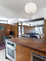 A post-renovation view of the kitchen shows it opening into the family room. Replacing the former white cabinets are Island drawer fronts and wall panelling of teak wood and reclaimed American elm countertop, milled by Vintage Material Supply. The differing grains of the teak veneers and elm countertop vadd complexity and rhythm to the kitchen's wood motif. Stuc Pierre plaster ceiling selected by the homeowner, Sloan Houser, adds an airy feel to the opened space.