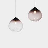 The Parison Pendant is a hand-blown glass pendant that mixes black and clear glass to create an ombre effect. Designed by Nat Cheshire, the top of the pendant is an opaque black that moves in a fluid gradient to ultimately become transparent. The neck of the pendant holds an LED lamp that diffuses soft, warm light. Nearly two feet in diameter, this overhead fixture creates a sculptural statement.
