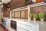 Instead of a typical backsplash above the oven, the designer opted for a row of windows to bring in additional natural light. The cooktop hood is sealed under cabinets to create a clean, streamlined effect, while custom tiles in modern shapes add texture. The oven, cooktop, and steam oven are Wolf. The dishwasher is Bosch.  Search “backsplash” from Bright Kitchen Remodel Contrasts Bright White and Dark Wood