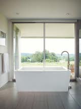 A crisp, white freestanding bathtub is the centerpiece of this modern bathroom, with a minimalist yet dramatic faucet. Views of the landscape out the window give the feeling of being outside while staying inside, and a retractable blind provides privacy when necessary. The floor-to-ceiling windows provide ample lighting for an open effect.