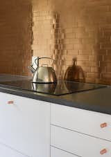 In the kitchen, copper-colored stainless steel tiles from TileBar create a glowing backsplash that is offset by the neutral white cabinets. The cabinetry is by IKEA, the countertop is Caesarstone, and the induction cooktop is by Bosch.