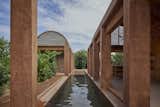 A long, narrow reflecting pool divides the rectilinear structures. Stained concrete pillars blend with clay-colored bricks, given their particular shade from prior use during the clay firing process at a local art studio.