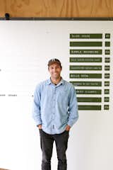 George Wilkins stands in front of a green chalkboard sign system that serves as a “to do” list for the business.  Photo 3 of 14 in This New Zealand Design Duo’s Headquarters Are as Fresh and Lighthearted as They Are