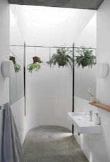 In the en suite bath on the second floor, a concrete floor gives way to a round, tiled, double-height space that culminates in a skylight. Plants hang on either side of a custom shower-head from Still Bathrooms. The faucets is from Crestial and the pendants are from Spazio Lighting.