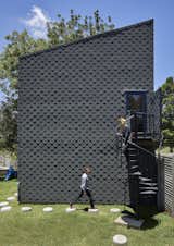 The modest two-story studio building by architect Greg Katz is made of a lightweight steel framework entirely clad in what is traditionally a roofing material: an asphalt-colored shingle, made of only two-millimeter-thick recycled rubber sheets, finished with a silicate coating (with a 20-year lifespan). The circular pavers are not actual pavers, but the residue from the pouring of the coffered slabs for House Katz. Instead of letting it go to waste, Katz asked the builders to pour the small amount of concrete left over from each newly mixed batch into a circular container. Once set, these circular shapes were popped out and stored to ultimately become a playful walkway.