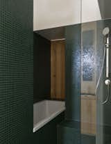 The bathroom is clad in moss-green tile, and a wooden shutter opens to a view of the living room fireplace.