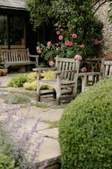Stone tile and wooden chairs in the backyard are surrounded by flowers, bushes, and trees.&nbsp;