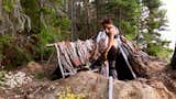 a structure of wood and bark being built by a woman in the middle of a dug pit