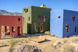 The property comprises three monolithic volumes: a green structure with the kitchen and living/dining room, a connected blue building with the bedroom and bath, and a freestanding pink gazebo that holds a long wooden table for covered outdoor dining.  Photo 4 of 7 in One Night in Joshua Tree’s Multicolored, Cubist Monument House