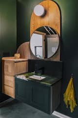 Bathroom with walls painted in dark green, wall-mounted sink, dark quartzite counter and backsplash, slate tile, curving elm wood vanity, circular mirror and wall-mounted light. 