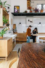 Women speaking at wood worktable in double-height kitchen with pedestal farm sink and white porcelain tile backsplash. 