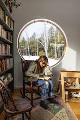 Girl sits in antique rocking chair reading a book by bookcase and round window.