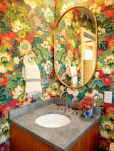 Bathroom with floral wallpaper, curving mirror, and under-mounted sink set into stone countertop with stone backsplash. 
