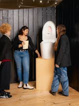 Group of women examine toilet with integrated bidet set upon a pedestal display in showroom.