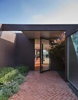 A wide brick patio leads to the main entrance, where a black-framed glass door opens to the sprawling, sunlit interiors.