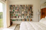 Bedroom with Rakks shelves housing book collection, whit walls, light wood floors, down comforter and view of grassy field