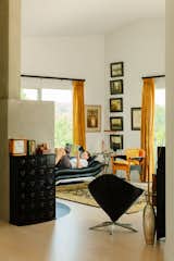 Man lays on black leather sofa reading a magazine in living room with white walls, light wood floors abstract art, hanging photographs, orange-and-black chair, black chair, black cabinet orange curtains, and black-and-gold floral rug