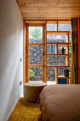 Bedroom with geometric shelving in front of large window looking out over side yard, yellow rug, ottoman, white walls, and a wood paneled ceiling
