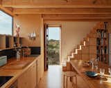 Wood paneled kitchen at sunset with stairs and in-built storage, exposed beams, and view of seaside hills through front door 