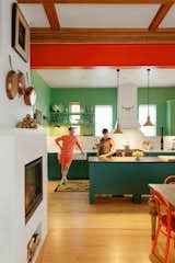 Jonathan Solomon and Meg Gustafson stand in kitchen of their postmodern pomo renovation of 19th century Uptown Chicago home with dark green cabinets, white countertops and backsplash, red painted steel I-beam, long brass pendant lamps, light green walls, shelves, and cabinets, light wood floors, and black metal fireplace framed in white venetian plaster.