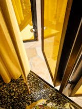 Detail of tiled patio shifting to black, white, brown, and beige terrazzo flooring across threshold of blackened steel-framed folding golden glass doors lined with golden curtains in detached office of Hollywood home.
