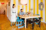 Kiki Goti stands beside Vincent Staropoli who sits at light wood dining table in their Clinton Hill, Brooklyn, New York City, loft apartment with white-and-yellow striped painted walls, blue and dark brown chairs, light wood floors, pendant lamp, OO+II metal-winged wall lamp, and mirror with striped black-and-white and orange, yellow, and blue frame.