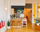 Kiki Goti and Vincent Staropoli cook in the kitchen of their Clinton Hill, Brooklyn, New York City, loft apartment with painted blue wall, high wooden stools, rolling wood sideboard, white countertops, light wood cabinets, OO+II metal-winged wall lamps, and white metal pendant lamp.