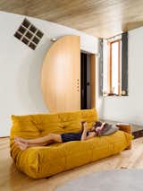 Girl lies on mustard sofa in living room of house in Libson, Portugal, with light wood floors, grey carpet, large, semicircular door, diamond glass block window, wood block side table, and curving ceiling.