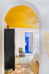 Bedroom of renovated home in Merida, Mexico, with rounded archway, peeling white walls, yellow ceiling, black armoire, black Sol chair by Chuch Estudio, faded colorful geometric and floral tiled floors, and yellow rug.