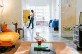 Small white dog looks on as architect Aranza Garcia searches through light wood cabinet decorated with black circles in living room of renovated home in Merida, Mexico, with decorative green and floral tiles, peeling walls, orange sofa and rug, black tables and desk, Chuch Estudio chairs, and glass paneled doors framed in metal painted pale pink. 