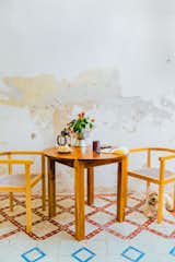 Small white dog sits beneath light wood chairs with ratan seating stand on white tiling with red and blue geometric designs next to peeling white wall and round table made of medium-toned wood in kitchen of renovated home in Merida, Mexico. 