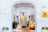 View of living room through archway in renovated home in Merida, Mexico, with peeling white walls, floral orange and green floor tiles, orange rug and sofa, black coffee tables and desk, and red spherical pendant light.