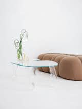Clara Jorisch’s glass coffee table around plants and brown plush sofa in white room in Montreal workshop.
