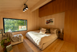 Loft bedroom of MOR.II prefab small home cabin by CABN with large grey bed, medium hard wood floors, large window, white sofa chair, exposed mass timber beam, and wood millwork storage