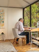 Man works at minimal, white metal framed desk and linen upholstered chair in room with large glass windows, white rug, and medium toned wood floors in home  in Camp Everhappy on San Juan Islands, Washington, restored by Jason F. McLennan.