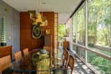Dining room with glass dining table supported on brass triangular pedestal, geometric brass pendant lamp, wood paneled walls, blue wallpaper with golden figures of trees, floor-to-ceiling glass windows, and slate floors midcentury internationalist home in Wyomissing, Pennsylvania by Muhlenberg Brothers and renovated by Kevin Yoder and Louise Cohen.