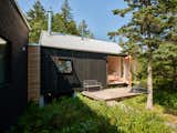 Home by Andrew Frederick in Owls Head, Maine, with black vertical wood siding, wood balcony outside bedroom with large open glass door, metal patio furniture, metal roof, and wild garden full of shrubs and trees.
