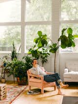 Man sits in pink velvet arm chair beneath tall windows in living room with white sofa, pink and white rug, plants in copper pots, and short curvy sculpture in living room of midcentury home in Columbus, Ohio, renovated by GRA+D, Greg Dutton of Midland architecture and Ashley Sargent Price.