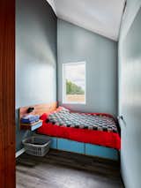 Bedroom with blue green grey walls with plywood platform bed, long wood headboard, light periwinkle blue shelves, drawers, and cubbies, and red and green quilt coverlet in The Ben Smith Welcome Home Center in New Orleans, Louisiana, by OJT.