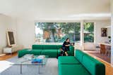 Woman sits working next to dog in living room in Philip Johnson Wolf House in Newburgh, New York, renovated by Jiminie Ha and Jeremy Parker with Hay green fabric sofas and chairs, large long windows overlooking forest, glass Barcelona Table by Mies van der Rohe with books, metal cabinetry by USM Modular, medium tone wood floors, dark red maroon tiling, and large wood table with mesh metal chairs.
