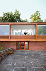 Couple lean on wood rail of balcony porch in Philip Johnson Wolf House in Newburgh, New York, renovated by Jiminie Ha and Jeremy Parker with brick first floor and wood clad second story with tall, wide, large windows, and slate paved front yard driveway.