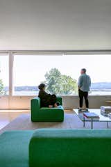 Living room in Philip Johnson Wolf House in Newburgh, New York, renovated by Jiminie Ha and Jeremy Parker with Hay green fabric sofas and chairs, large long windows overlooking lake, mountains, and forests, and glass Barcelona Table by Mies van der Rohe with books.