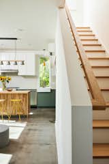 Staircase with wood tread and railing leading up from kitchen with wooden island, yellow stools, white and dark turquoise cabinetry, pendant lighting, white apron sink, and polished concrete floors.