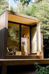 Woman stands in floor-to-ceiling and black metal-framed aux box model 110 prefab prefabricated home with wood deck and black painted steel cladding.