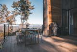 Wood table with chairs and bench and gas grill sit outside Cedar-sided, glulam prefab prefabricated home by Blend Projects with large deck with glass-paneled railing in British Columbia, Canada.