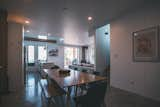 Dining room with long wood table, black chairs, and bench, white MDF and plywood wall panels, and double-height kitchen, living room, and deck in background in cedar-sided, glulam prefab prefabricated home by Blend Projects in British Columbia, Canada.