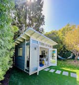 Studio Shed's prefab prefabricated Signature studio model with swinging glass doors, grey blue fiber cement lap siding, and white shed roof in backyard.