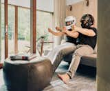 Couple wearing racing helmets sits on grey couch pretending to drive race cars in den with grey and white carpet and dark grey leather pouf chair in house in Palisades New York by Charles P Winter renovated by Rick Cook Fox CookFox.